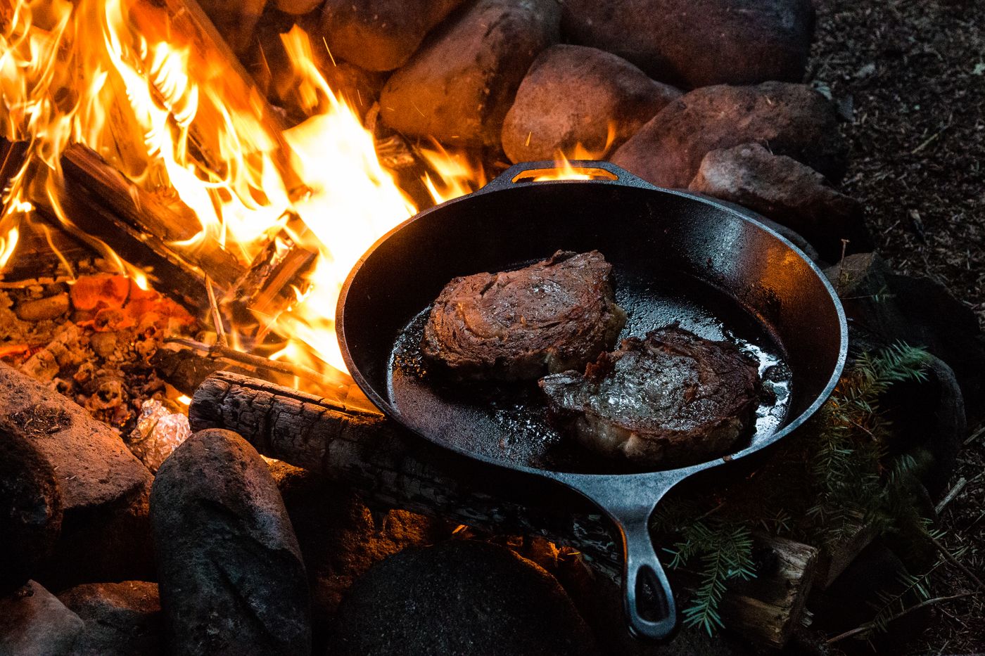 A cast-iron pan of food cooking on an open fire among stones and logs